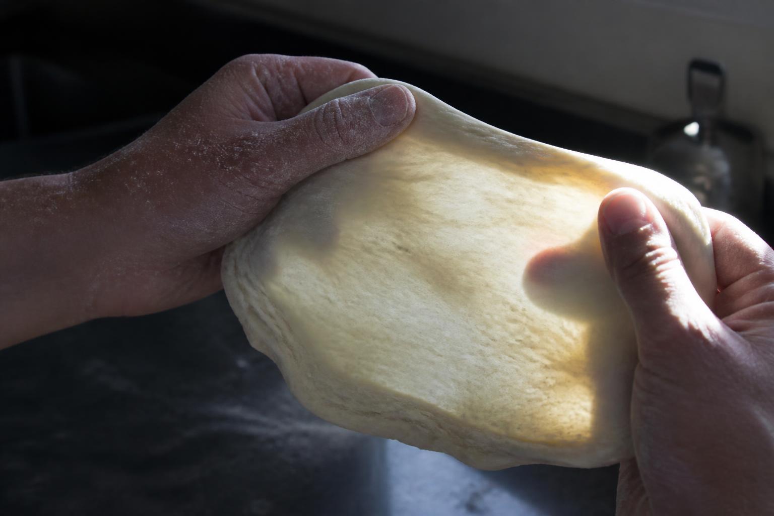 Hands stretching pizza dough into a thin translucent membrane against daylight