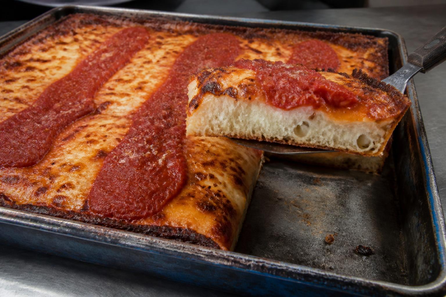 Detroit-Style-Pizza im blauen Stahlblech mit karamellisiertem Käserand und zwei Streifen Tomatensauce, ein Stück angehoben