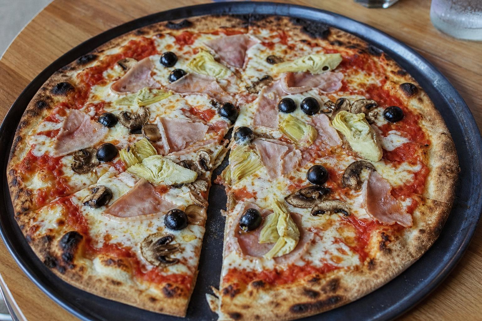 Dünne, knusprige Pizza Tonda Romana als Capricciosa mit Kochschinken, Artischocken, Champignons und Oliven auf dunklem Blech